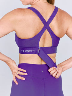A woman shown from the back wearing a bright purple Ultimate Sports Bra and matching leggings, with the brand name visible on the waistband. The woman is cinching the back ribcage band.