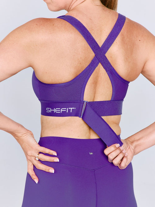 A woman shown from the back wearing a bright purple Ultimate Sports Bra and matching leggings, with the brand name visible on the waistband. The woman is cinching the back ribcage band.