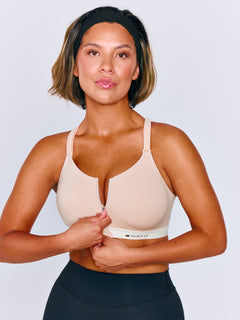 A woman wearing a beige sports bra with a white underband and a front zipper, shown against a light gray studio background. The brand name 'SHEFIT' is visible on the band.