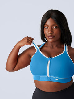 A woman modeling a bright blue Flex Sports Bra with white trim and a front zipper, paired with black leggings, against a light gray studio background. The woman is lifting the shoulder strap of the bra.