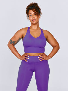 A woman modeling a matching purple athletic set consisting of an Emerge Sports Bra and CloudNine Legging, posed against a light gray studio background.