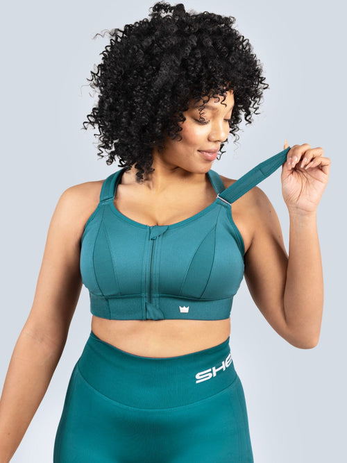 Woman demonstrating the adjustable shoulder strap on a teal SHEFIT sports bra.
