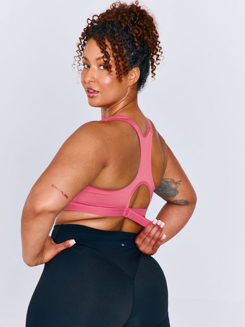 A woman modeling a bright pink Emerge Sports Bra from the back, showing the open back design with adjustable ribcage band straps, paired with black leggings against a light gray studio background.