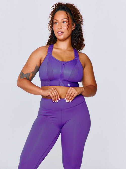 A woman modeling a matching bright purple athletic set consisting of Ultimate Sports Bra and CloudNine Legging photographed against a solid light gray studio background.