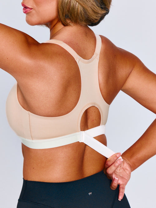 Rear view of a woman wearing a beige sports bra with a white underband and adjustable strap, shown against a light gray studio background. Woman is adjusting the ribcage band on the sports bra.
