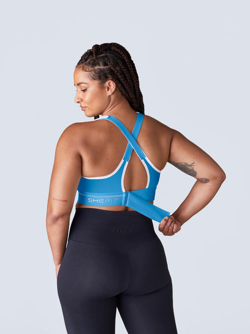 A woman shown from the back wearing a bright blue Flex Sports Bra with white trim paired with black leggings, against a light gray studio background. The woman is cinching the back ribcage band of the bra.