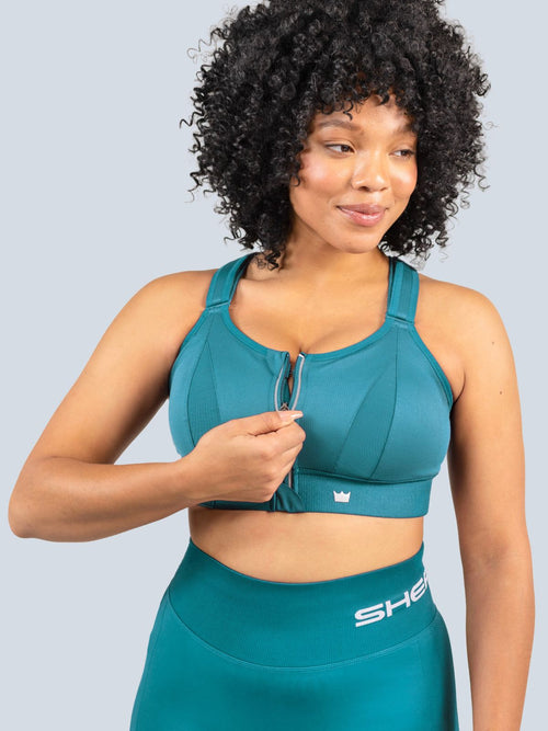 Woman wearing a Focused Teal Ultimate Sports Bra and Focused Teal Seamless Shorts with a visible brand logo on a light gray background. Zipping the front zipper of the bra.