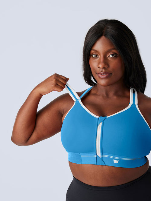 A woman modeling a bright blue Flex Sports Bra with white trim and a front zipper, paired with black leggings, against a light gray studio background. The woman is lifting the shoulder strap of the bra.