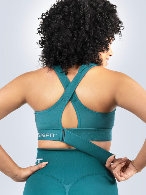 Woman wearing a Focused Teal Ultimate Sports Bra and Focused Teal Seamless Shorts with a visible brand logo on a light gray background. Cinching ribcage band.