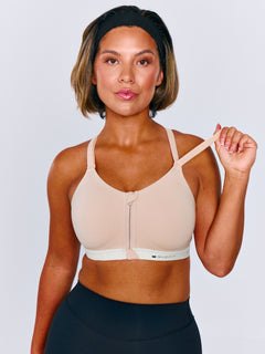 A woman modeling a beige sports bra with a white underband with a front zipper and thin adjustable straps, paired with black leggings, against a light gray studio background. Woman is adjusting the shoulder strap on the sports bra.