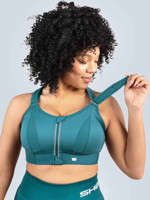 Woman wearing a Focused Teal Ultimate Sports Bra and Focused Teal Seamless Shorts with a visible brand logo on a light gray background. Lifting one shoulder strap.