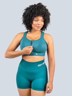 Woman holding the front zipper of a teal SHEFIT sports bra, paired with matching shorts.