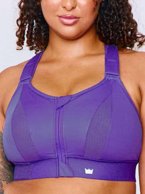 A close-up of a woman wearing a bright purple Ultimate Sports Bra with a front zipper and a small white crown logo on the lower right side, set against a light gray background.