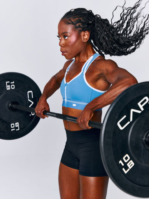 A woman posing in a bright blue Flex Sports Bra with white trim and black leggings while lifting, set against a light gray studio background.