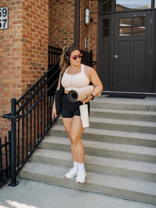 A woman in activewear stands on outdoor concrete stairs in front of a brick building, holding a rolled yoga mat and a white tumbler. She is wearing a beige sports bra with a white underband, black shorts, white socks, and white sneakers.