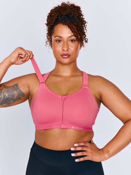 A woman modeling a bright pink Emerge Sports Bra with a front zipper and wide shoulder straps, paired with black athletic bottoms, against a light gray studio background. The woman is lifting the shoulder strap of the sports bra.