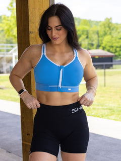 A woman wearing a bright blue Flex Sports Bra with white trim and black high-waisted shorts with white branding, standing outdoors under a wooden pavilion.