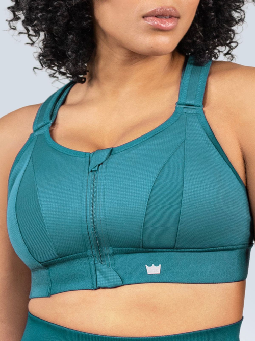 Close-up of a teal SHEFIT sports bra showing textured fabric, seamed cups, and front zipper.